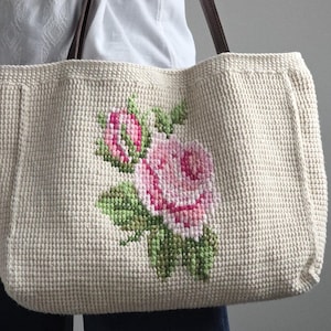 Könnte beinhalten: Cremefarbene Tragetasche mit einem Kreuzstich-Rosenmuster in Rosa und Grün. Die Tasche hat braune Ledergriffe. Die Tasche besteht aus einem gewebten Material.