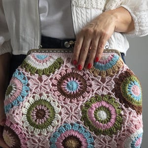 Könnte beinhalten: Handgefertigte Häkeltasche mit Metallverschluss. Die Tasche hat ein farbenfrohes Blumenmuster in Rosa, Blau, Grün und Braun. Die Tasche wird von einer Person gehalten, die eine weiße Jacke und blaue Jeans trägt.