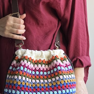 Könnte beinhalten: Eine mehrfarbige Häkeltasche mit cremefarbenem Oberteil und braunem Lederriemen. Die Tasche hat ein lebendiges Muster aus rosa, orangefarbenen, blauen und grünen Quadraten. Die Tasche wird von einer Person getragen, die ein weinrotes Hemd trägt.