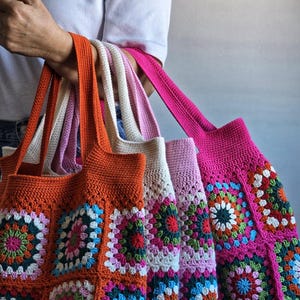 May include: Three crocheted tote bags with colorful square patterns. The bags are made with different colored yarn, including orange, pink, and white. The bags are stacked on top of each other, with the orange bag on the bottom, the pink bag in the middle, and the white bag on top.