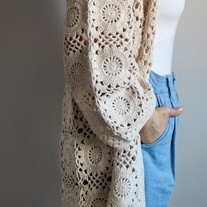 Könnte beinhalten: Beige Häkel-Cardigan mit offenem Frontdesign und floralem Muster. Das langärmlige Kleidungsstück besteht aus einem weichen, offen gewebten Material. Der Cardigan wird über einem weißen Oberteil und einer blauen Jeans getragen.