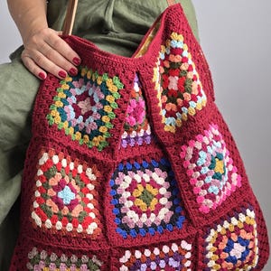 May include: A large, burgundy crochet tote bag with a patchwork design. The bag features colorful, square granny squares in various shades. It has brown leather straps and a spacious interior, perfect for carrying essentials.