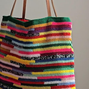 Può includere: Una borsa tote realizzata in filato lavorato all'uncinetto multicolore con un motivo a righe orizzontali nei toni del giallo, rosa, verde, blu, rosso e viola. La borsa presenta due sottili manici in pelle liscia color cuoio.