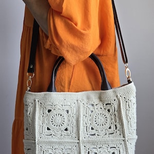 Könnte beinhalten: Cremefarbene Häkeltasche mit floralem Muster. Die Tasche hat einen braunen Ledergriff und einen langen, verstellbaren Riemen. Die Tasche wird von einer Person getragen, die ein orangefarbenes Kleid trägt.