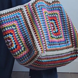 May include: A large, colorful crochet tote bag with a white border and black leather handles. The bag is made with a variety of different colored yarns, creating a vibrant and eye-catching design.
