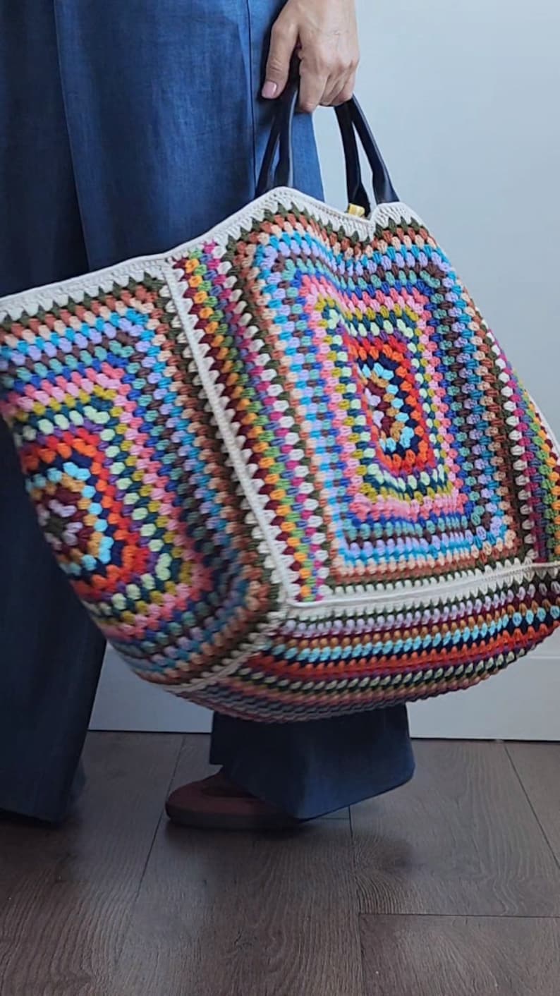 May include: A large, colorful crochet tote bag with a white border and black leather handles. The bag is made with a variety of different colored yarns, creating a vibrant and eye-catching design.