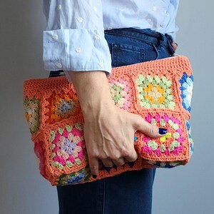 Crochet zipper clutch bag with bright multicolor granny-square panels and coral edging, held by a person in a light blue shirt.