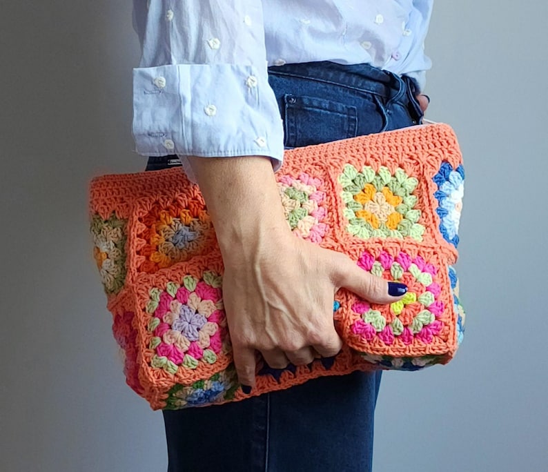 Crochet zipper clutch bag with bright multicolor granny-square panels and coral edging, held by a person in a light blue shirt.