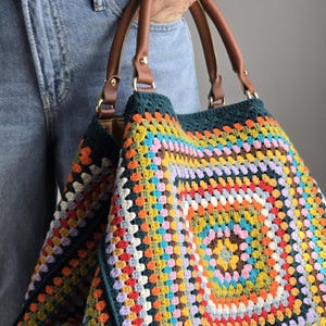 May include: A colorful, handmade crochet tote bag with a granny square design. The bag features a vibrant mix of colors, including blue, yellow, orange, and green. It has brown leather handles and gold-colored hardware.