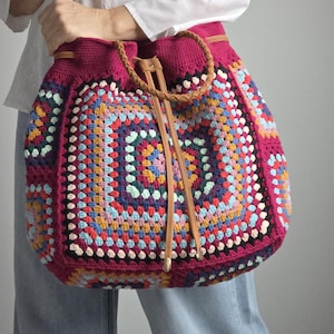 Könnte beinhalten: Handgefertigte gehäkelte Tragetasche mit einem lebendigen Granny-Square-Muster. Die Tasche hat eine tiefmagenta Basis mit bunten Quadraten in Blau-, Gelb- und Grüntönen. Sie hat einen braunen Lederriemen und einen Kordelzugverschluss.