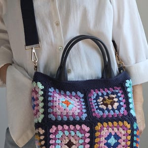 Könnte beinhalten: Eine marineblaue Häkeltasche mit einem farbenfrohen Granny-Square-Design. Die Tasche hat einen schwarzen Lederaufsatz und -griffe sowie einen schwarzen, verstellbaren Schulterriemen mit goldfarbener Hardware. Es ist ein handgefertigter Artikel.