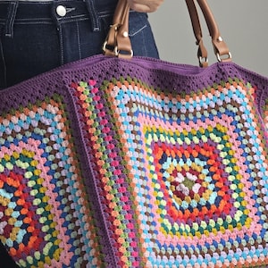 May include: A large tote bag featuring a multi-colored granny square crochet pattern with a purple border. The bag includes two brown leather handles attached with gold-toned metal hardware and a solid purple crocheted base and side panels.