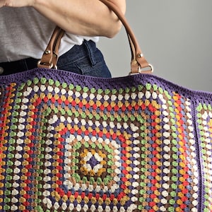 Könnte beinhalten: Große, farbenfrohe Häkeltasche mit Granny-Square-Design. Die Tasche hat einen lila Rand und braune Ledergriffe. Das Häkelmuster umfasst verschiedene Farben wie Grün, Rot, Gelb und Weiß.