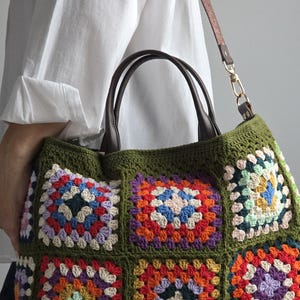 May include: A handmade, olive green crochet tote bag with a colorful granny square design. The bag features a brown leather handle and a detachable brown strap. The granny squares are in various colors, including red, orange, blue, and purple.