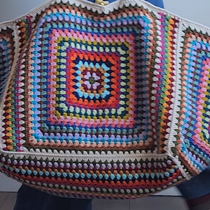 May include: A large, colorful crochet tote bag with a square pattern in the center. The bag is made with a variety of colors, including red, orange, yellow, green, blue, purple, and brown.
