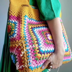 May include: A handmade crochet tote bag in a granny square pattern. The bag features a mustard yellow base and straps, with a colorful patchwork design in pink, blue, green, and orange. A small leather tag reads "Handmade".