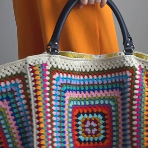 May include: A large, colorful crochet tote bag with a black leather handle. The bag is made of a variety of different colored yarns, including red, orange, yellow, green, blue, purple, and pink. The bag has a large, square design in the center, surrounded by a border of smaller squares.