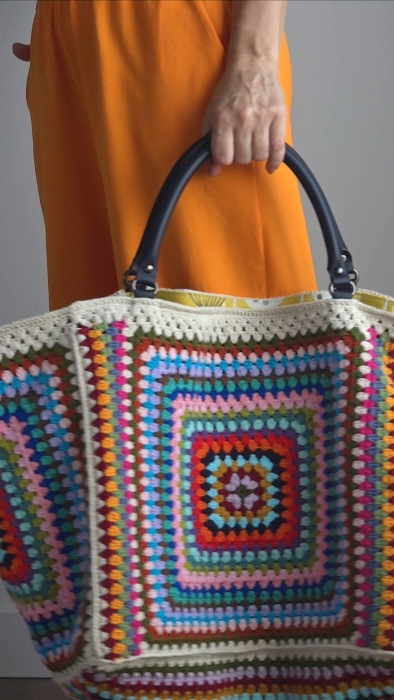 May include: A large, colorful crochet tote bag with a black leather handle. The bag is made of a variety of different colored yarns, including red, orange, yellow, green, blue, purple, and pink. The bag has a large, square design in the center, surrounded by a border of smaller squares.
