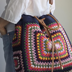 Może przedstawiać: Duża, ręcznie robiona torba z szydełka z kolorowym wzorem granny square. Torba ma granatową podstawę i jest ozdobiona kwadratami w różnych kolorach, w tym różowym, pomarańczowym, żółtym i zielonym. Posiada brązowe skórzane paski i zamknięcie na sznurek.
