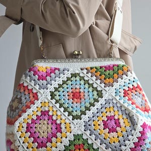 Könnte beinhalten: Eine Häkeltasche aus mehrfarbigen Granny Squares in den Farben Pink, Gelb, Grün, Blau und Weiß. Die Tasche verfügt über einen metallischen Schnappverschluss mit geprägtem Muster und einen breiten, cremeweißen Schulterriemen aus Stoff.