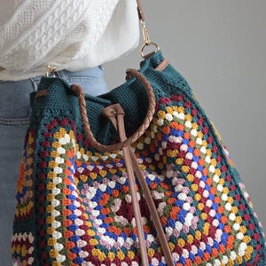 Könnte beinhalten: Große, handgefertigte Häkeltasche mit farbenfrohem Granny-Square-Design. Die Tasche hat einen tealgrünen Oberteil und Griff, mit einem braunen Lederriemen und Griff. Die Tasche ist ein stilvolles Accessoire.