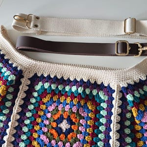 May include: A crocheted bag with a colorful granny square pattern. The bag has two straps, one brown leather and one white canvas.