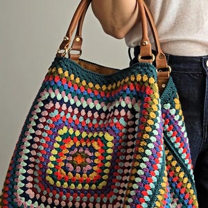 Könnte beinhalten: Eine große, farbenfrohe Häkeltasche mit einem quadratischen Muster in einer Vielzahl von Farben, darunter Rot, Blau, Gelb und Grün. Die Tasche hat braune Ledergriffe und goldfarbene Beschläge. Die Tasche ist etwa 46 cm breit und 38 cm hoch.