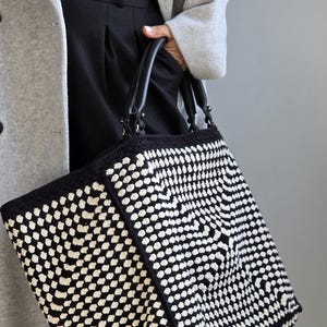 Può includere: Una grande borsa tote in crochet bianco e nero con manici in pelle nera. La borsa ha un motivo geometrico.