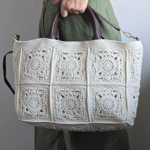 Può includere: Una borsa a mano in crochet con manico in pelle marrone. La borsa è realizzata in filato beige e ha un motivo quadrato.