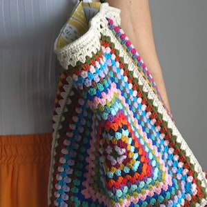 May include: A colorful crocheted tote bag with black leather handles. The bag is made with a granny square pattern and features a variety of colors, including red, orange, yellow, green, blue, purple, and pink.