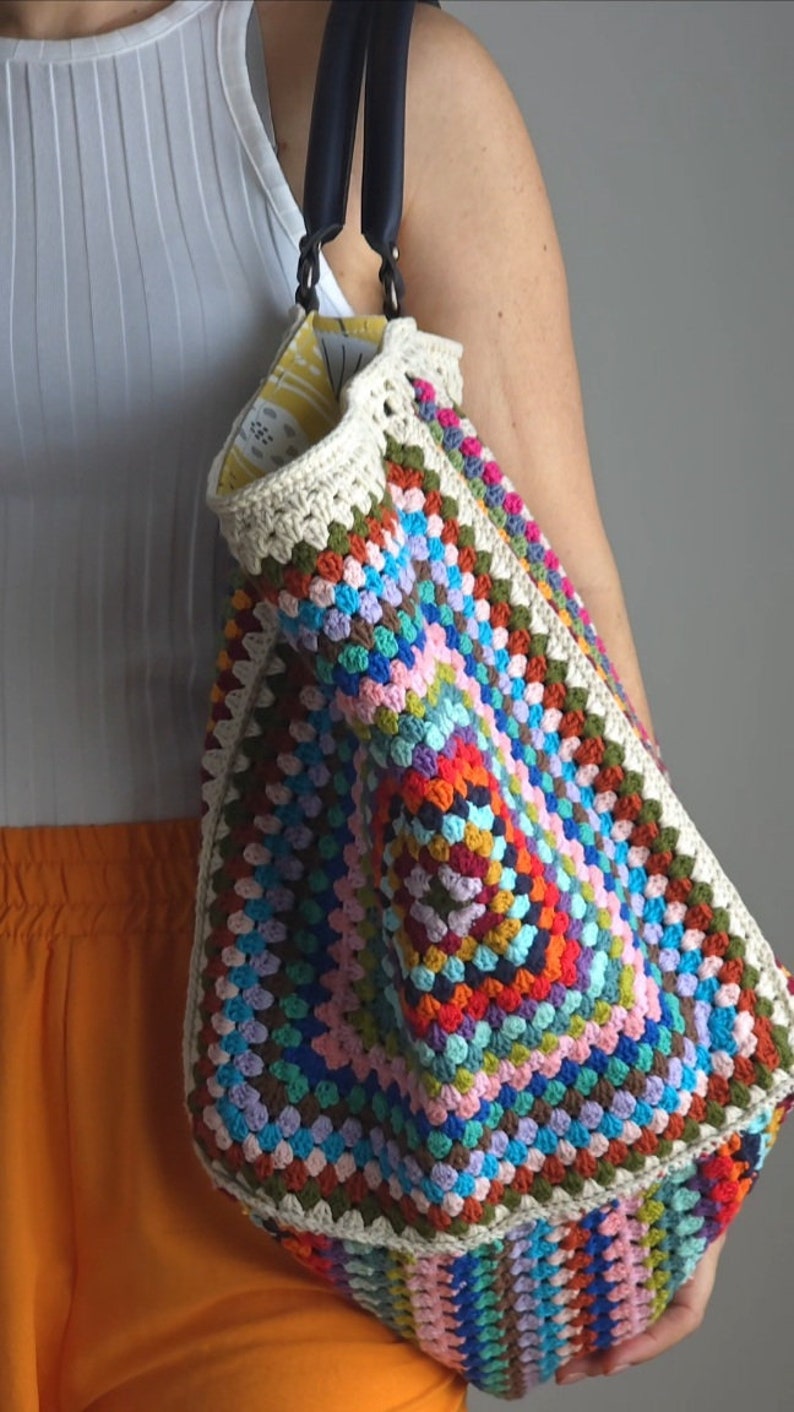 May include: A colorful crocheted tote bag with black leather handles. The bag is made with a granny square pattern and features a variety of colors, including red, orange, yellow, green, blue, purple, and pink.