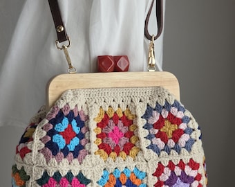 Puede incluir: Un bolso de ganchillo con un patrón de cuadrados de la abuela multicolor en tonos rosa, azul, amarillo y rojo, enmarcado por un asa superior de madera clara. El bolso incluye dos correas de cuero marrón oscuro y dos cierres rojos.