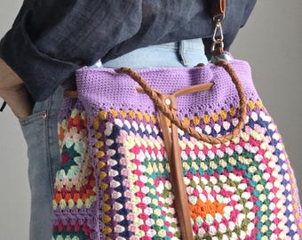 May include: A handmade crochet bag with a colorful granny square design. The bag features a purple top and a brown leather strap. The bag is a shoulder bag style.