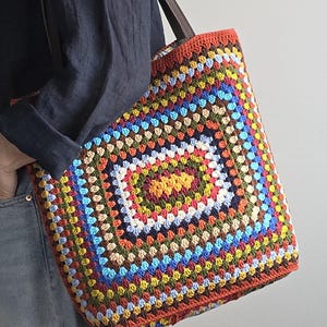 Può includere: Una borsa tote rettangolare all'uncinetto con un motivo a piastrelle granny multicolore nei toni del rosso, giallo, blu, verde e marrone. La borsa presenta un bordo all'uncinetto in tinta unita rosso-arancio sui bordi superiore e inferiore.