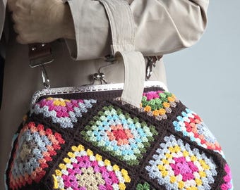Puede incluir: Un bolso de crochet colorido y hecho a mano con un cierre metálico. El bolso presenta un diseño de patchwork con cuadrados de varios colores, incluyendo rosa, amarillo, verde y azul, sobre un fondo marrón oscuro. Tiene un asa y una correa para el hombro.