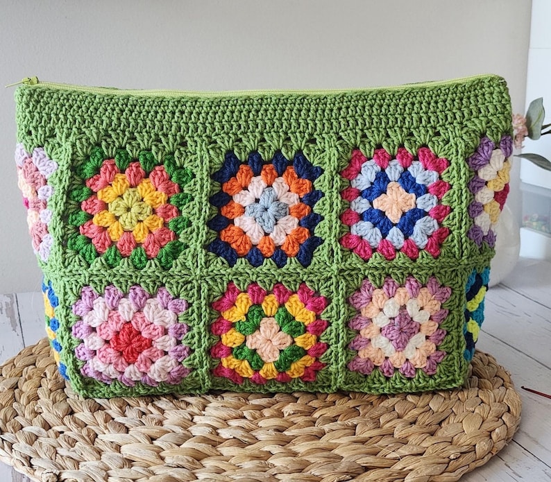 Crochet zipper pouch clutch bag with granny square patchwork in bright colors, green edging, top zipper, on a woven mat.