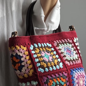Könnte beinhalten: Handgefertigte Tragetasche mit bordeauxroter Basis und bunten Häkelquadraten. Die Quadrate weisen verschiedene Farben wie Blau, Gelb, Rosa und Orange auf. Die Tasche hat einen schwarzen Griff und Lederakzente.