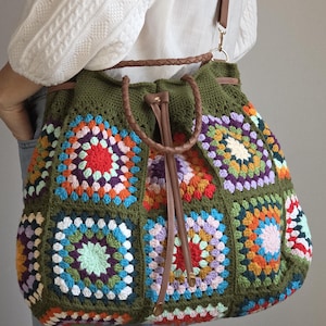 Könnte beinhalten: Eine große, olivgrüne Häkeltasche mit einem farbenfrohen Patchwork-Design. Die Tasche hat einen Kordelzugverschluss und braune Lederriemen. Die Patchwork-Quadrate sind in verschiedenen Farben gehalten, darunter Rot, Orange, Blau und Lila.
