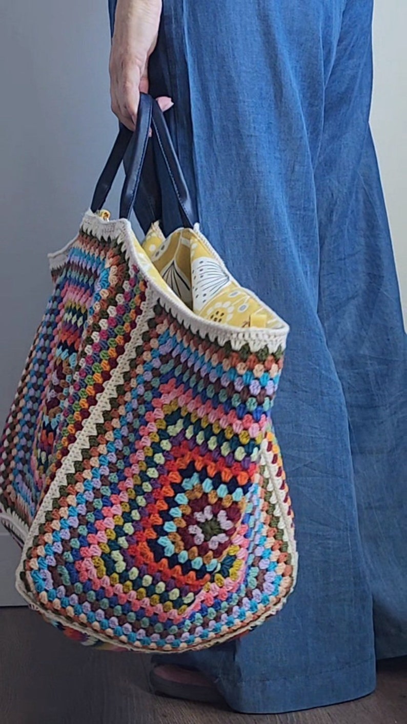 May include: A large, colorful crochet tote bag with a white lining and black handles. The bag is filled with a yellow and white patterned fabric.