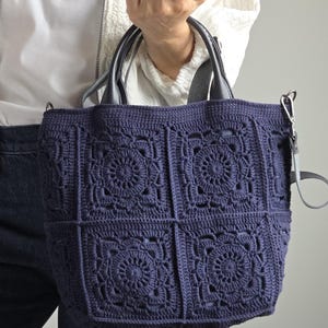 May include: A navy blue crochet tote bag with a floral pattern. The bag has black handles and a detachable shoulder strap. The bag is approximately 14 inches wide and 12 inches tall.