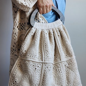 Könnte beinhalten: Eine beige gehäkelte Tragetasche mit einem Holzgriff. Die Tasche ist aus dickem, strukturiertem Garn gefertigt und hat eine quadratische Form mit abgerundeten Ecken.
