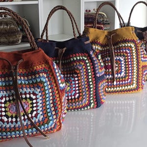 May include: A row of colorful crochet tote bags with brown leather handles. The bags feature a square granny stitch design in various colors, including red, blue, yellow, and green. The bags have a drawstring closure and are displayed on a white surface.