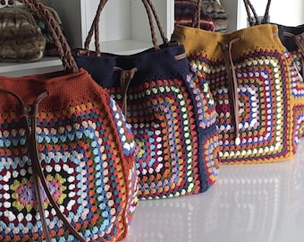 May include: A row of colorful crochet tote bags with brown leather handles. The bags feature a square granny stitch design in various colors, including red, blue, yellow, and green. The bags have a drawstring closure and are displayed on a white surface.