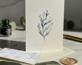 May include: A blank greeting card with a watercolor illustration of blue and gray seaweed. The card is on top of a cream-colored envelope, with a matching patterned insert. A gold wax seal and green ribbon are also present.