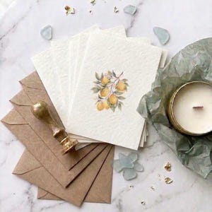 May include: A selection of stationery items, including cream cards with a lemon branch illustration, brown envelopes, a gold wax seal, and a candle in a gold tin. The items are arranged on a marble surface.