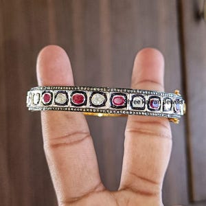 Puede incluir: Un brazalete rígido con bisagra que presenta una hilera de rubíes rojos ovalados y piedras blancas claras alternadas en un marco de metal oscuro. La superficie está incrustada con pequeñas piedras pavé y el texto Shree Kalyan Jewels es visible.