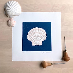 May include: A white and pink seashell print on a square of dark blue paper. Two real seashells and two carving tools are arranged around the print. The print is signed and numbered.