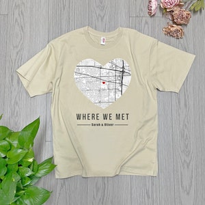 Puede incluir: Una camiseta beige con un mapa en blanco y negro de una ciudad en forma de corazón. El mapa tiene un corazón rojo en el centro. El texto "WHERE WE MET - Sarah & Oliver" está impreso debajo del corazón.