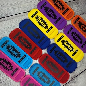 May include: A set of 11 colorful crayon-shaped fabric wristlets with personalized names in black lettering. The wristlets are arranged in a diagonal line on a wooden surface.