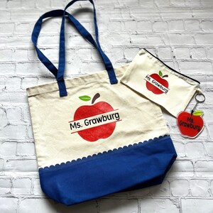 Könnte beinhalten: Ein Set Lehrergeschenke mit einem roten Apfeldesign mit dem Namen "Ms. Grawburg". Das Set beinhaltet eine Tragetasche mit blauen Henkeln und einem blauen Boden, eine kleine Reißverschlusstasche und einen Schlüsselanhänger.